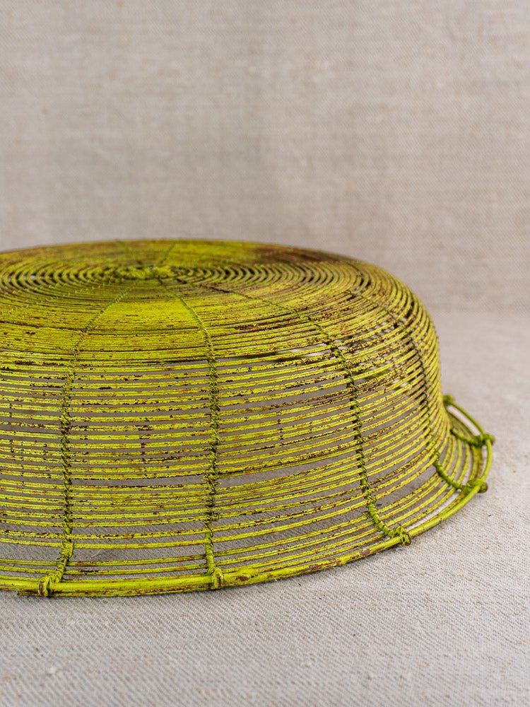 Antique French wire basket, green-painted fil de fer, upside down on beige textured surface.