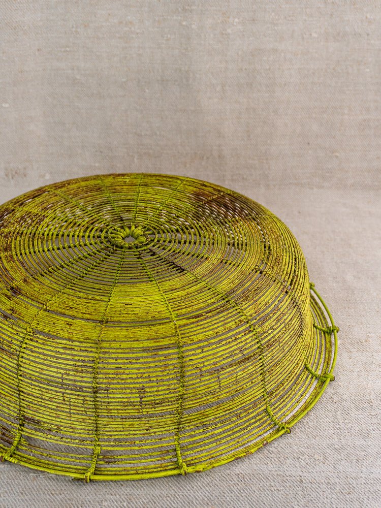 Antique French wire basket, yellow-green fil de fer, round mesh, gently worn, on beige textured surface.