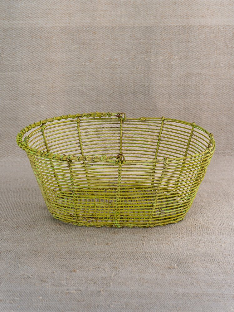 Antique French wire basket with green painted finish, oval form, on beige textured fabric.