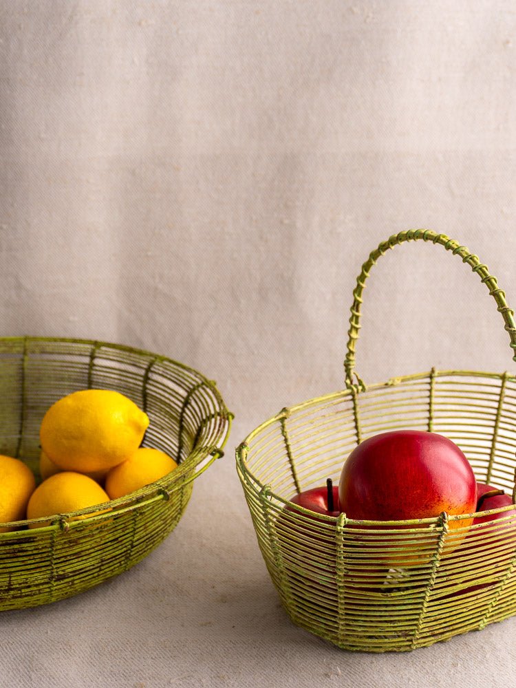 Pair of green-painted antique French wire baskets, one with lemons, one with apples, on fabric.