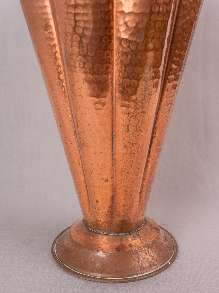Vintage French umbrella stand in hammered copper and brass with vertical ridges, flared base, on neutral fabric background.
