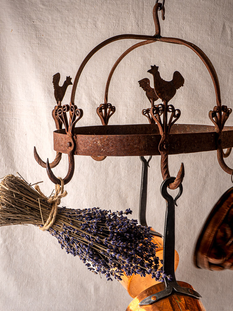 Vintage French wrought iron pot rack with rooster motif, utensils, and dried lavender on fabric background.