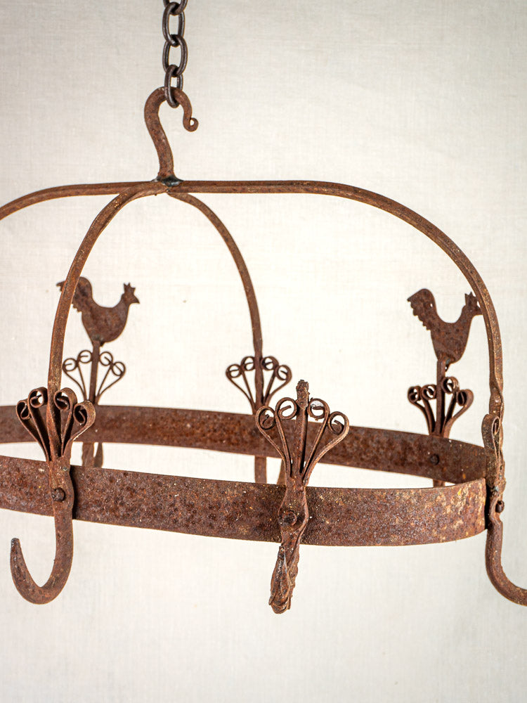 Vintage French wrought iron pot rack with rooster accents and hooks, chain-suspended, on light background.