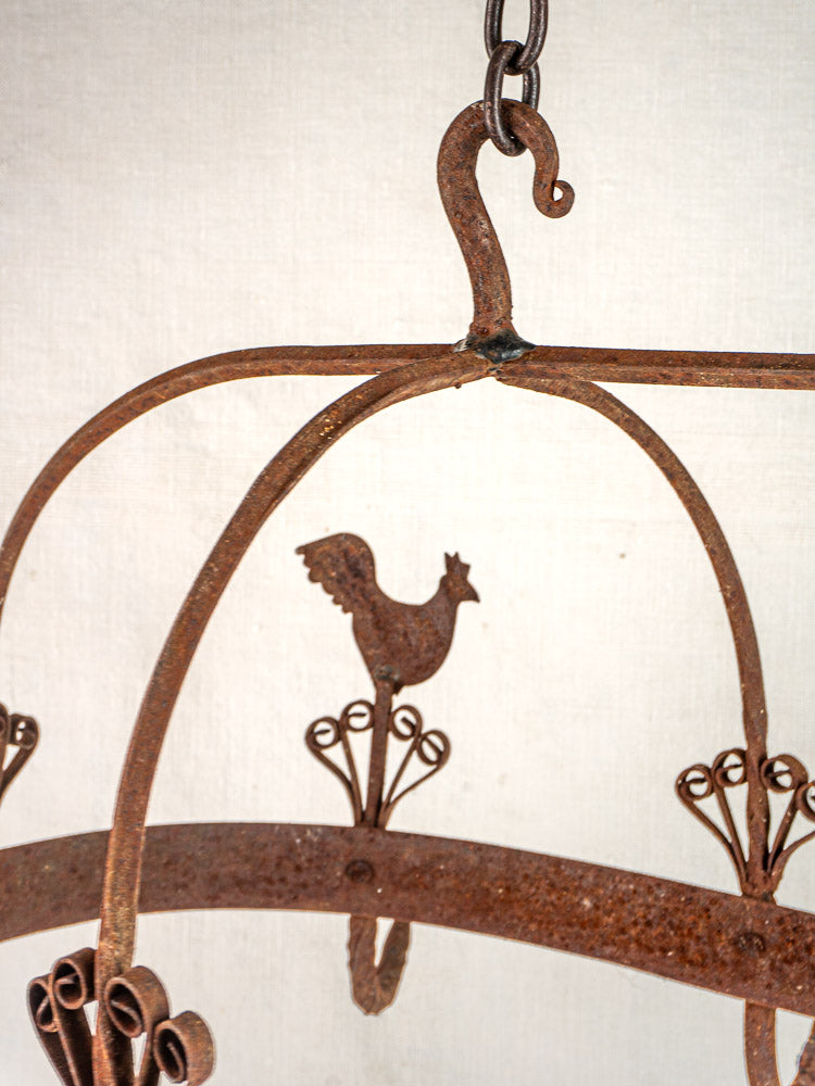 Vintage French wrought iron pot rack with rooster detail and scrollwork, hanging by chain on off-white background.