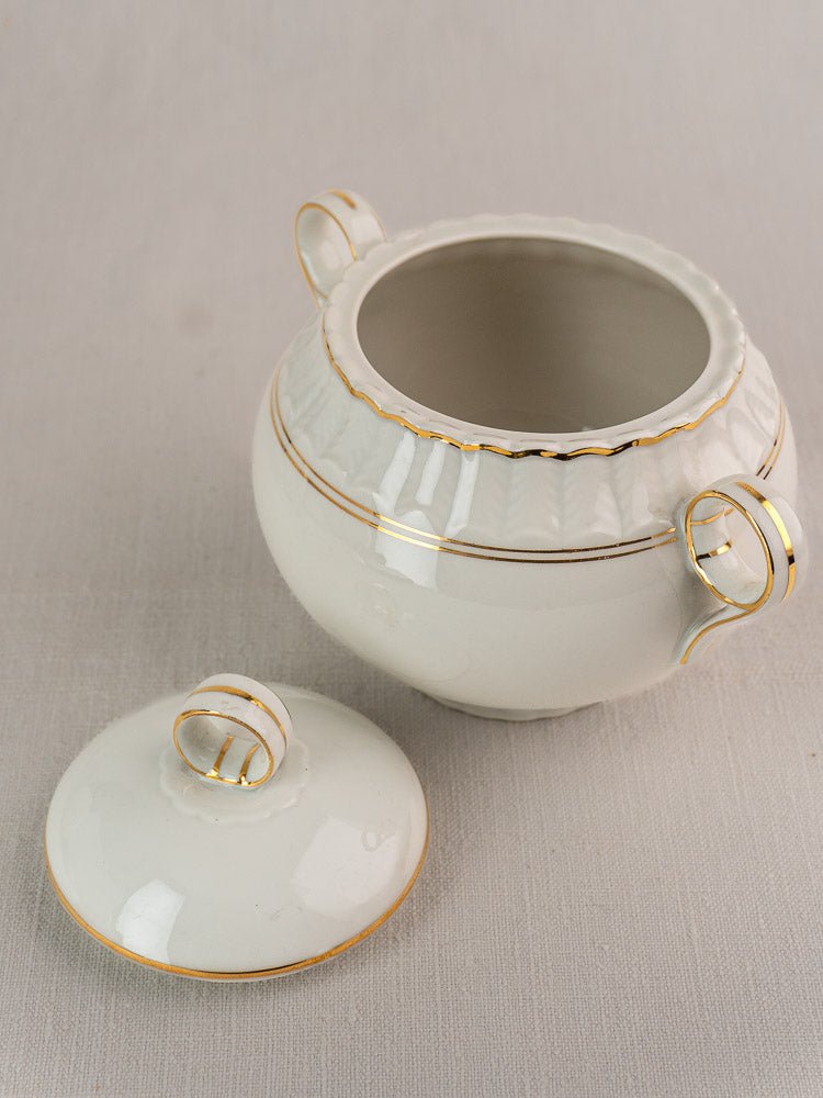 White Bernardaud porcelain sugar bowl with three gold-trimmed handles and separate lid on fabric.