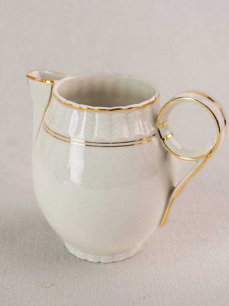 Antique French Limoges Bernardaud creamer, white porcelain with gold trim and scalloped rim on light surface.