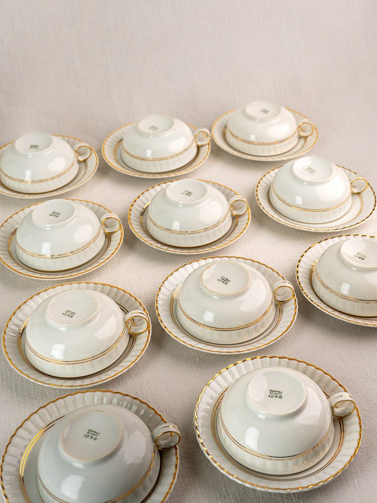 Twelve white Bernardaud Limoges teacups with saucers, gold trim, "Made in Japan" stamps visible.