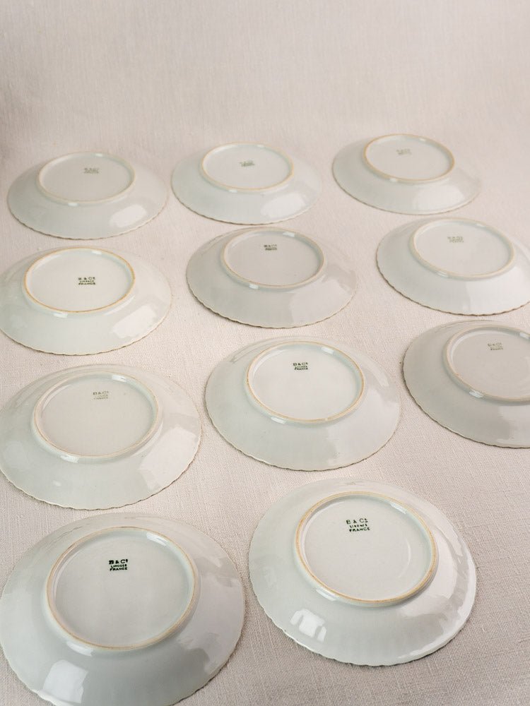 Twelve antique French Limoges Bernardaud porcelain saucers, gold-trimmed with green maker’s marks visible.