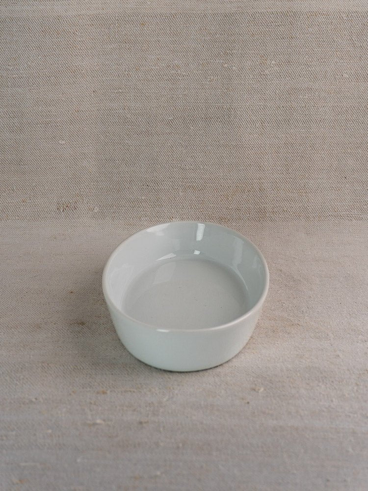 Small white ceramic bowl on textured beige fabric, reminiscent of vintage French porcelain serving ware.