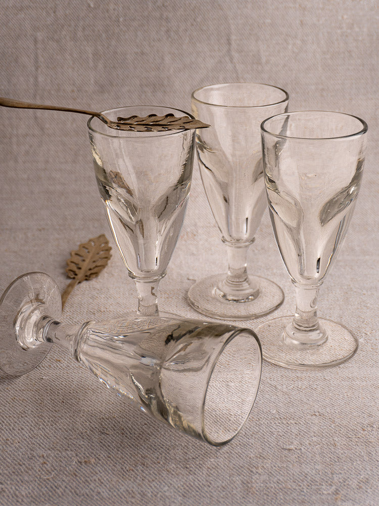 Set of four antique French uranium absinthe glasses with brass leaf spoon on beige textured surface.