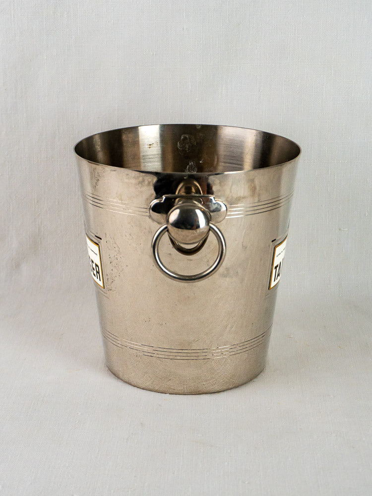 Taittinger vintage French plated wine cooler in stainless steel with front handle and side labels on a light gray surface.