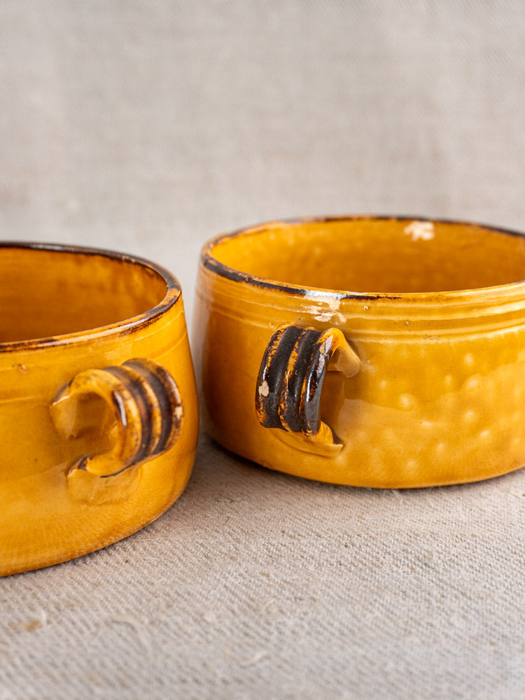 Pair of vintage yellow French Vallauris ceramic bowls with small handles and brown accents on a beige textured surface.