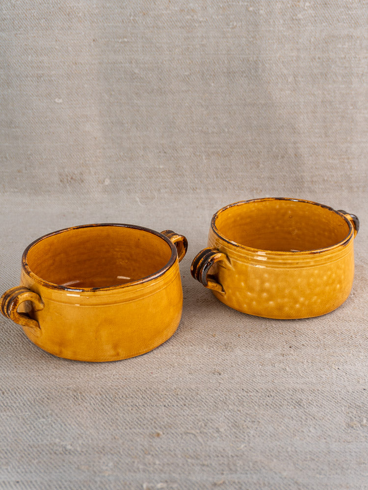 Pair of round yellow vintage French Vallauris bowls with side handles, shiny glaze, on textured beige fabric.