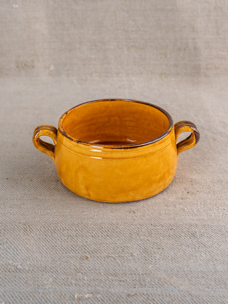 Glossy yellow vintage French Vallauris bowl with two handles on a beige textured fabric surface.