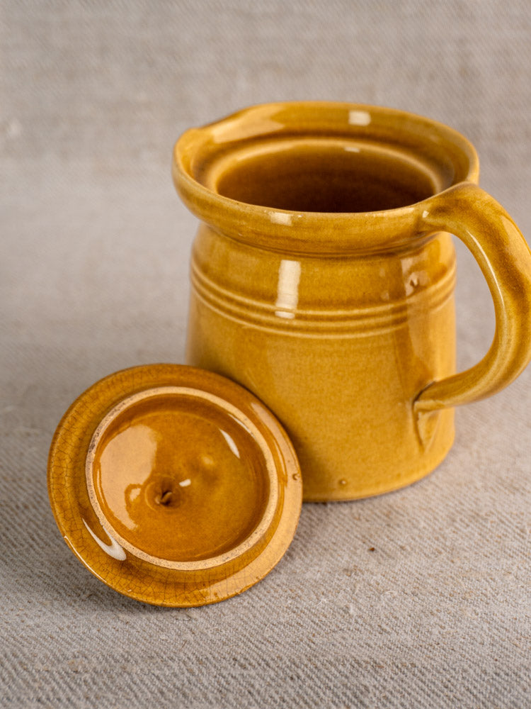 Antique French lidded pot à lait with honey ochre glaze on light gray textured fabric surface. Mid-20th century design.