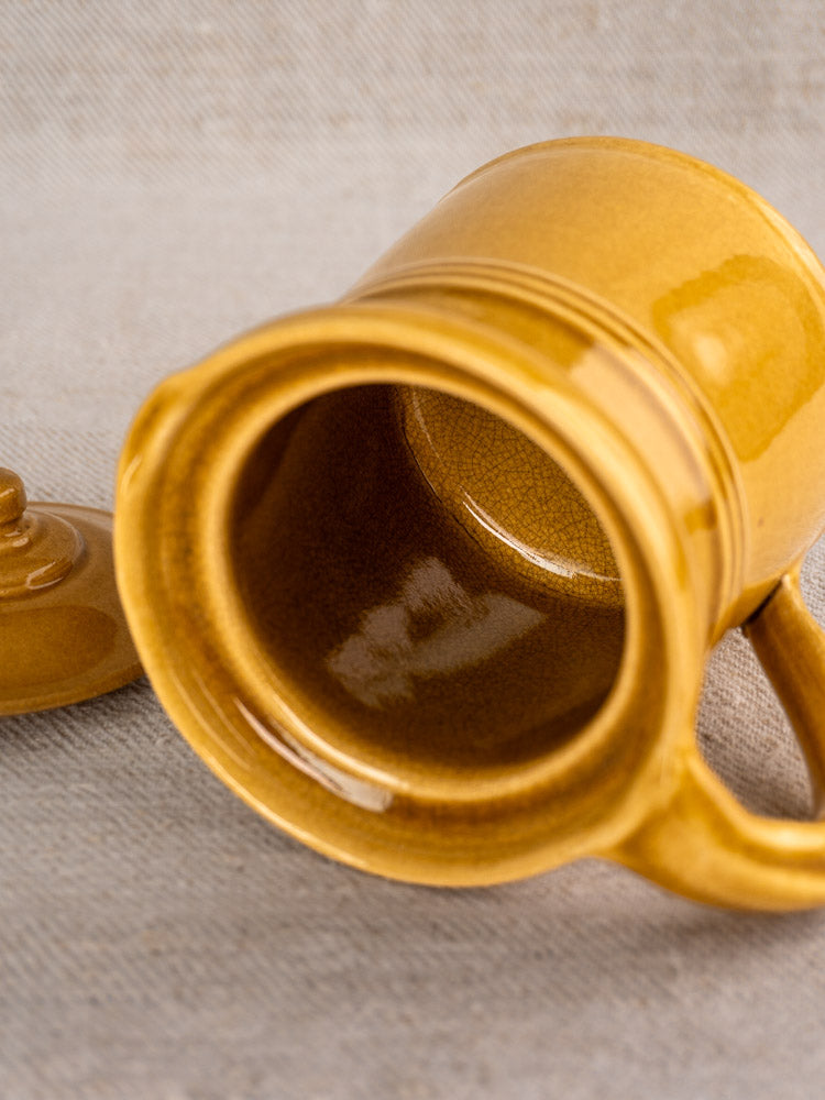 Mid-century French antique lidded pot à lait in honey ochre glaze, lid off, empty on textured beige surface.