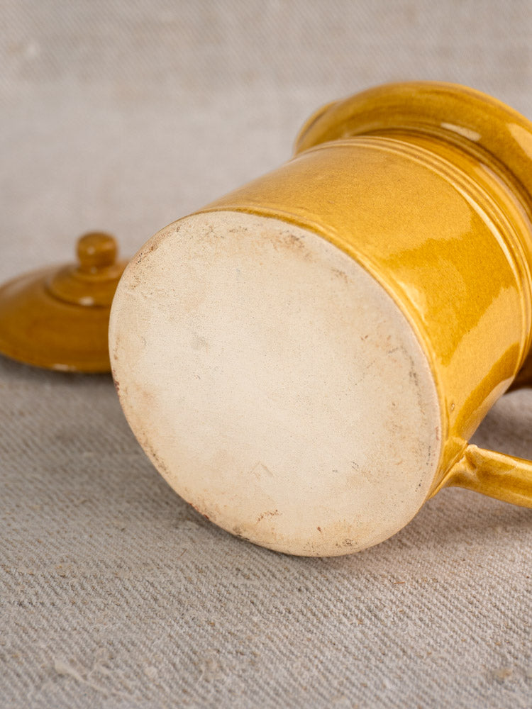Antique French lidded pot à lait with honey ochre glaze, unglazed base, and lid beside on beige textured surface.
