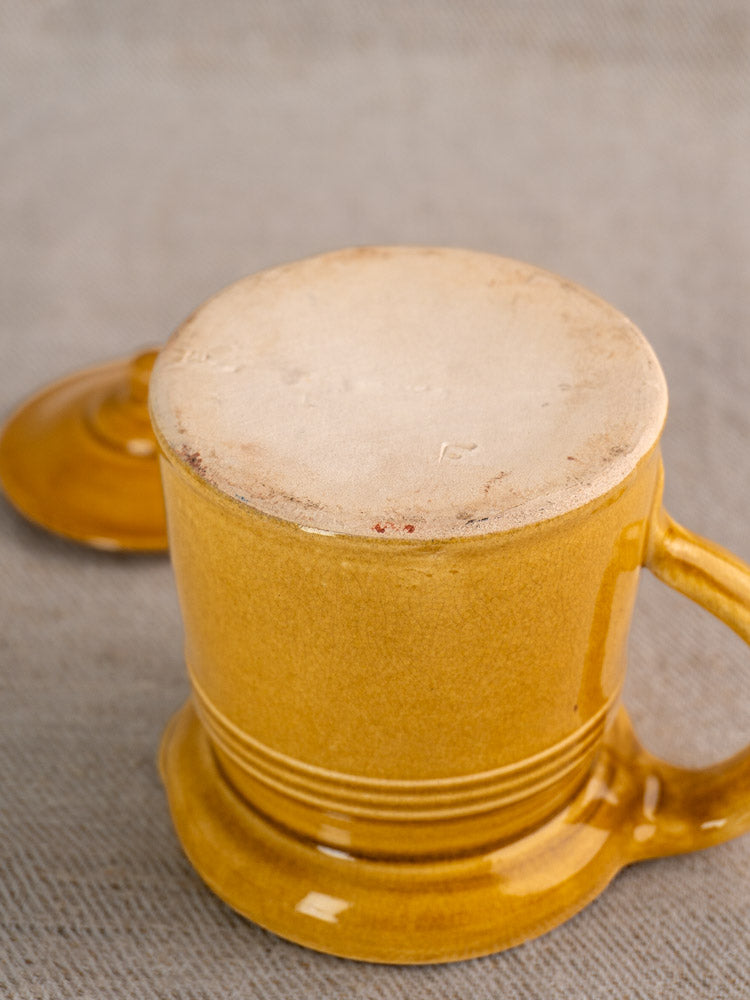 Antique French lidded pot à lait, honey ochre glaze, upside down showing unglazed base; lid rests on beige textured surface.