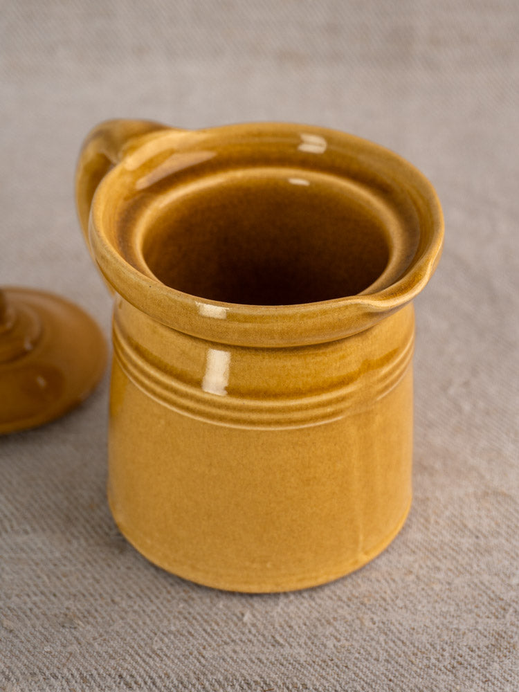 Antique French ceramic pot à lait with honey ochre glaze and lid on textured beige surface.