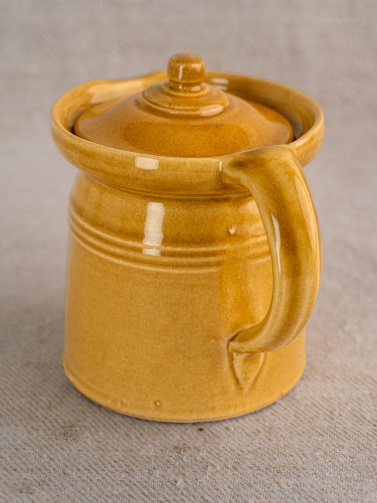 Antique French lidded pot à lait with honey ochre glaze and large handle on textured beige fabric.