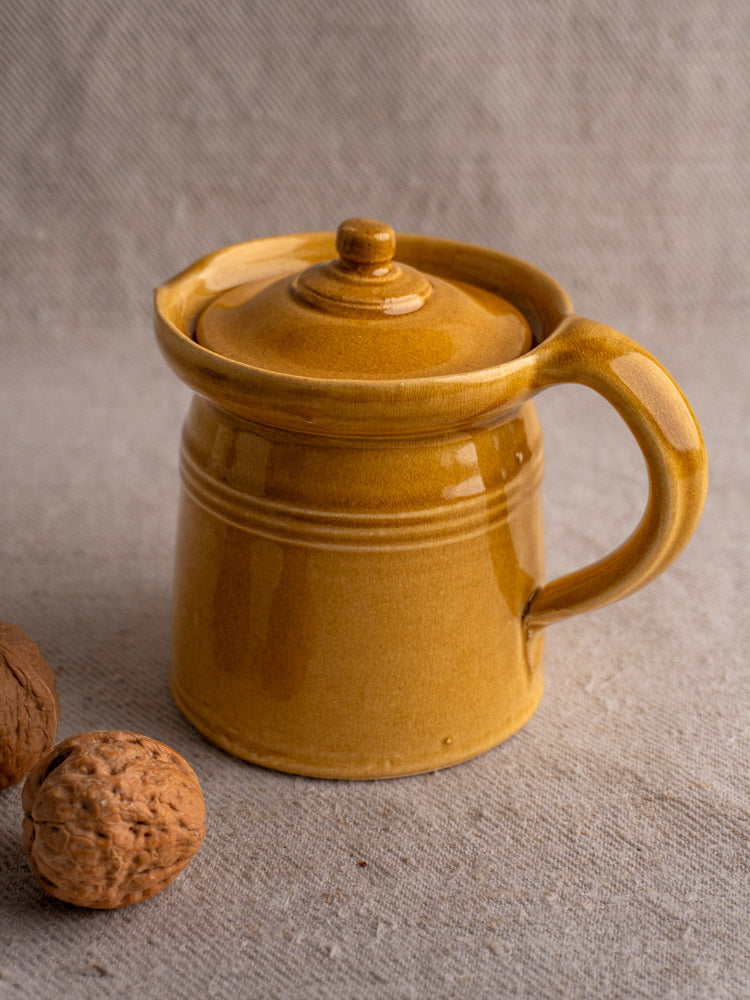 Antique French lidded pot à lait with handle and honey ochre glaze, on light fabric with walnuts. Mid 20th century.