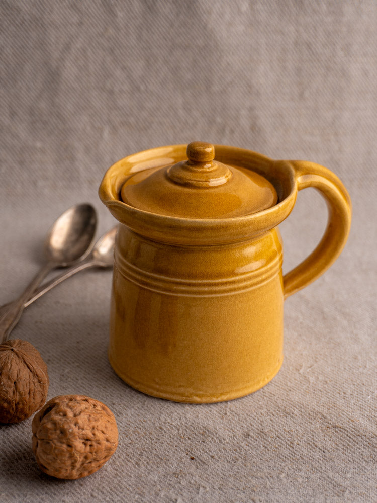 Antique French lidded pot à lait with honey ochre glaze, mid 20th century, on textured fabric with walnuts and silver spoons.
