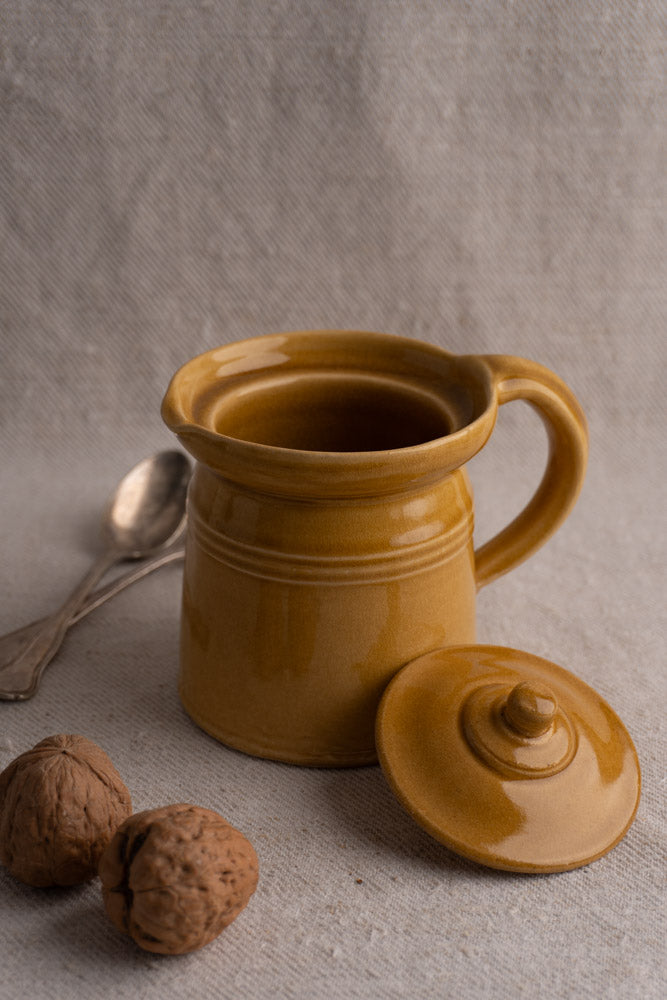 Antique French lidded pot à lait in honey ochre glaze with lid, walnuts, and vintage silver spoons on beige textured fabric.