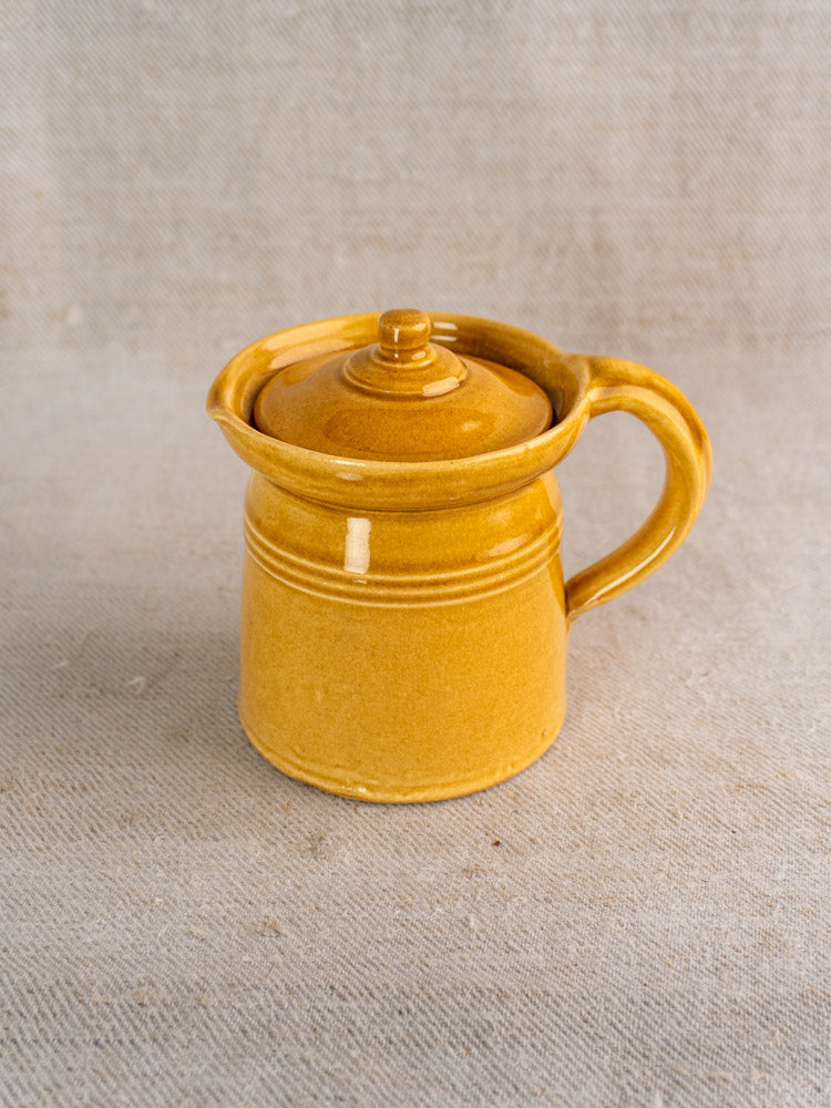 Antique French lidded pot à lait with honey ochre glaze and handle, set on textured beige fabric background.