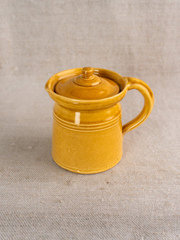 Antique French lidded pot à lait with honey ochre glaze and handle, set on textured beige fabric background.