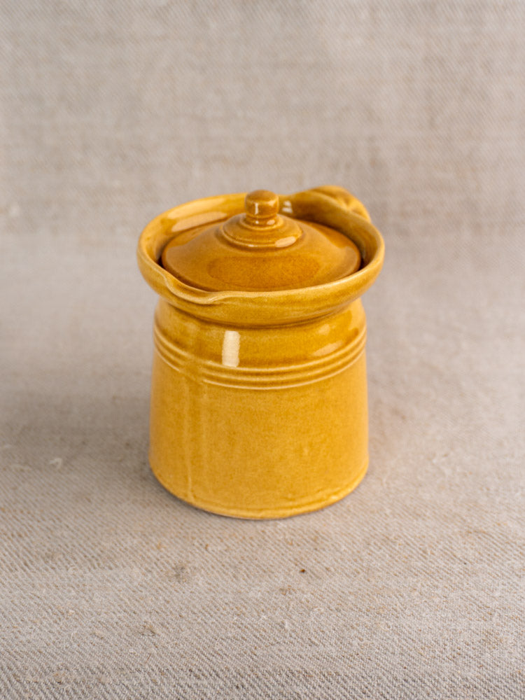 Antique French lidded pot à lait with honey ochre glaze and notched rim on beige textured fabric.