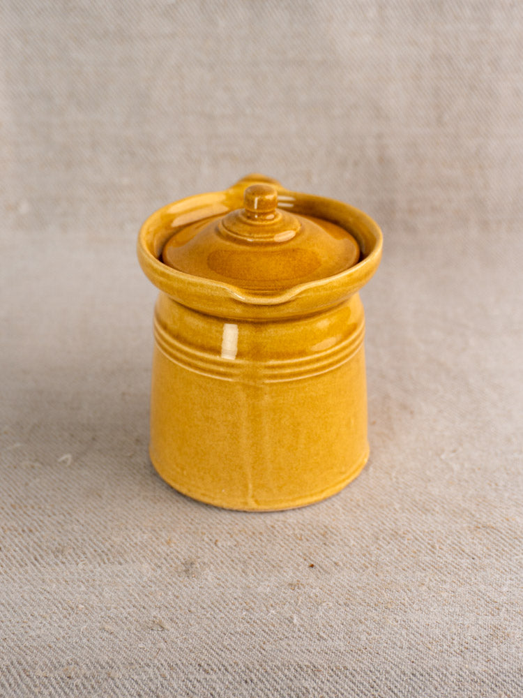 Antique French lidded pot à lait in honey ochre glaze with matching lid on textured beige fabric background.