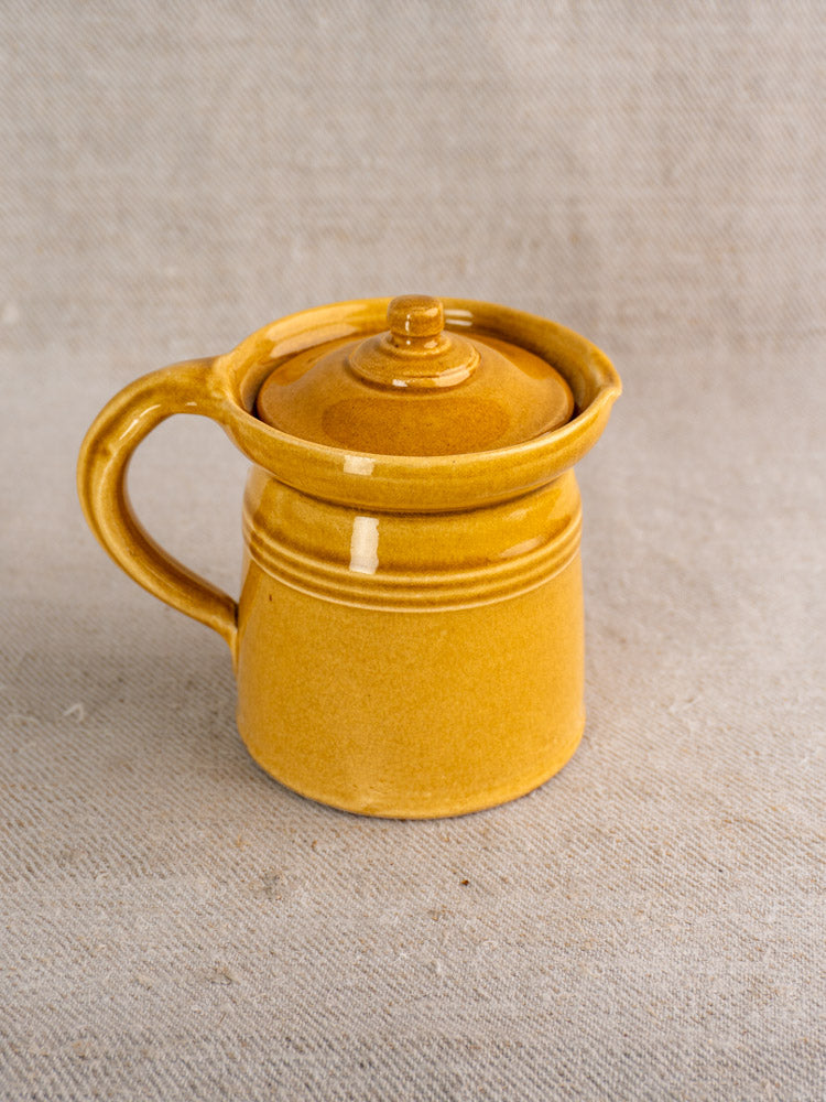 Antique French lidded pot à lait with honey ochre glaze and large handle on beige textured fabric background.