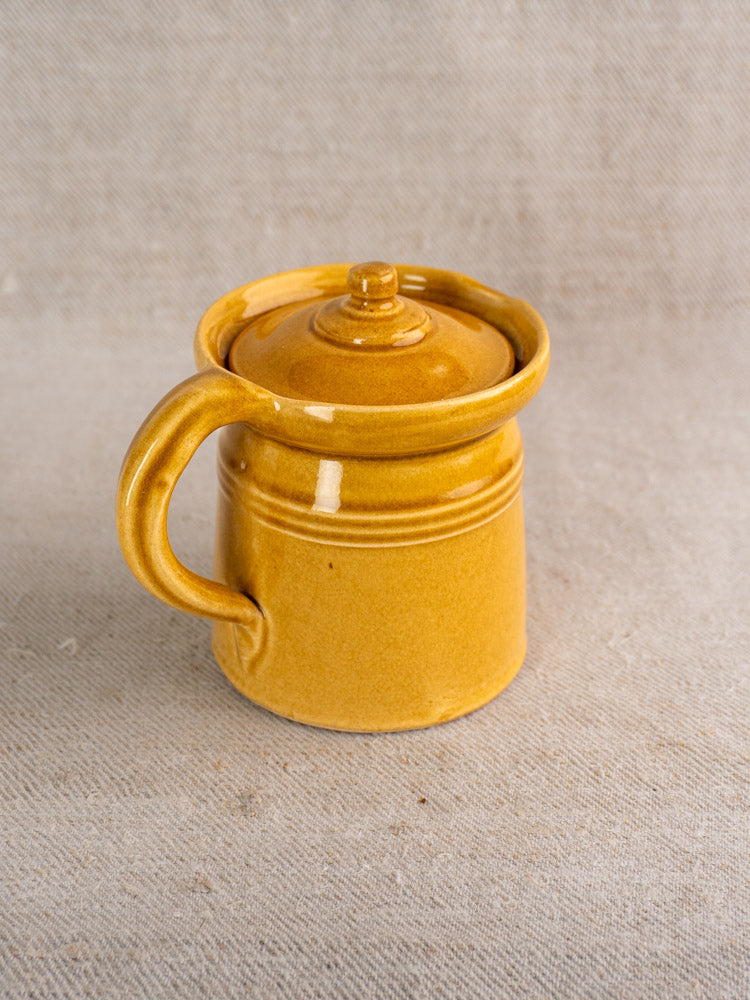 Antique French ceramic lidded pot à lait in honey ochre glaze with large handle on beige textured fabric.
