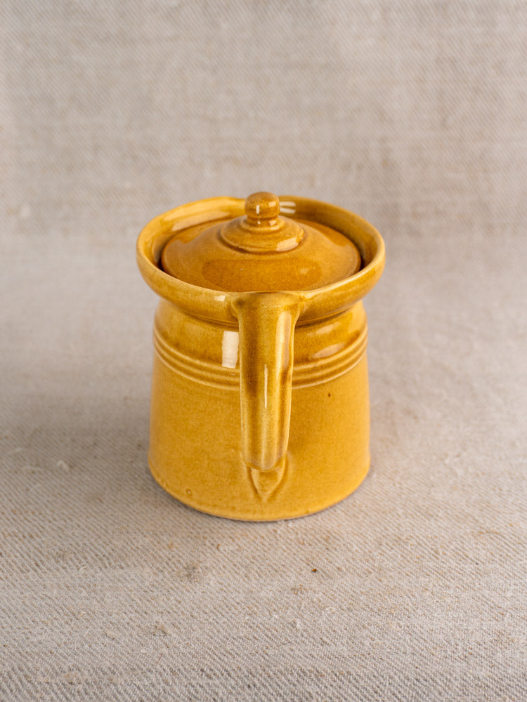 Antique French lidded pot à lait with honey ochre glaze and single handle on beige textured fabric.