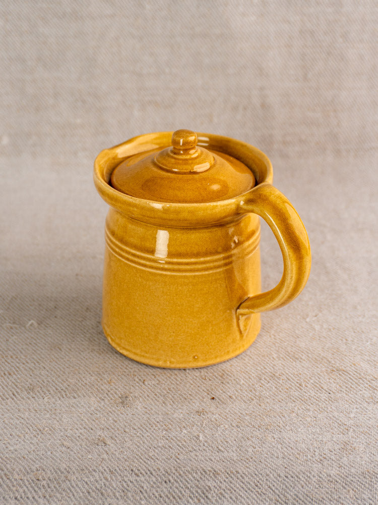 Antique French lidded pot à lait with honey ochre glaze and large handle on textured beige fabric.