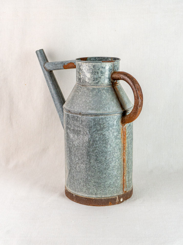 Vintage French galvanized zinc watering can with spout and rustic handle on a light fabric background.