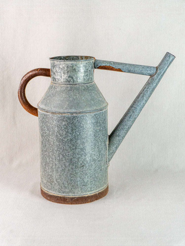 Vintage French galvanized zinc watering can with long spout and rounded handle, slight rust, on fabric.