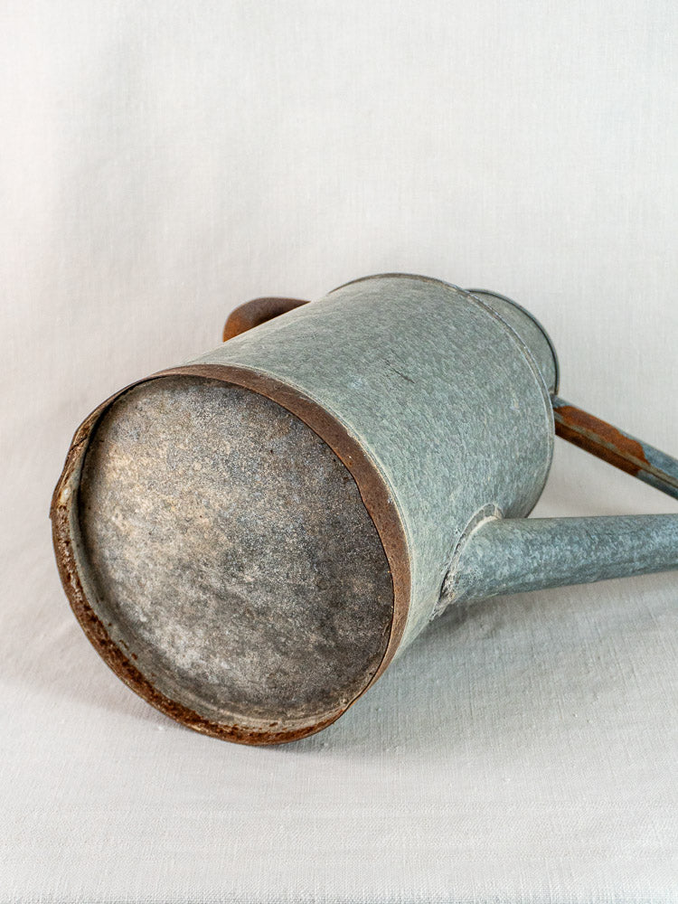 French galvanized zinc vintage watering can on white fabric, showing textured, timeworn bottom and angled spout.