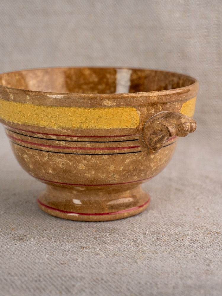 Antique French faience bowl by HB et Cie Choisy-le-Roi with yellow and red jaspé stripes, paw handle on beige fabric.