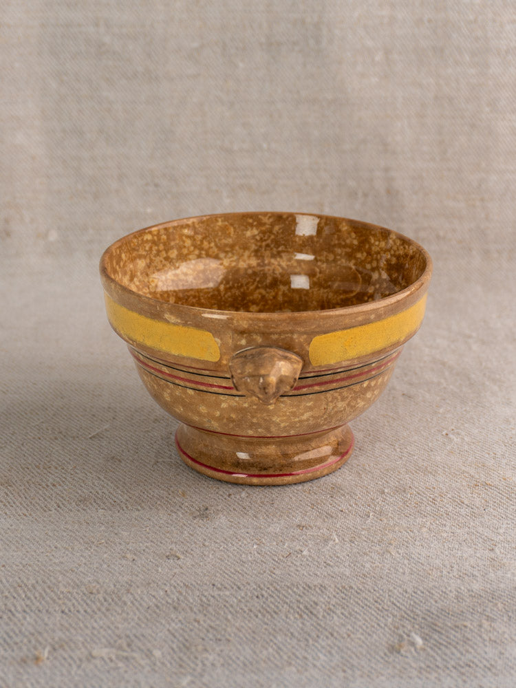 Antique French faïence bowl by HB et Cie Choisy-le-Roi with brown glaze, yellow stripe, red accents, lion head; on beige fabric.