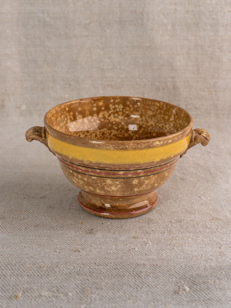Antique French faience bowl by HB et Cie with brown jaspé glaze, yellow rim, side handles on beige fabric background.