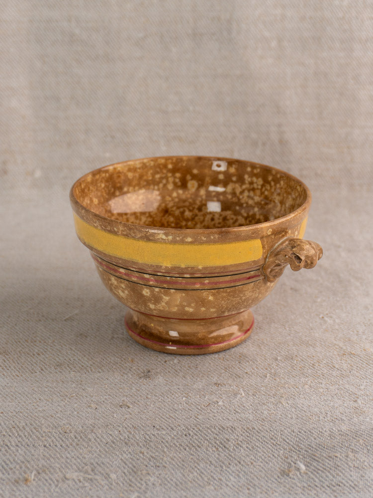 Antique French faience bowl by HB et Cie with mottled brown glaze, yellow rim, red accents, and handle on beige fabric.