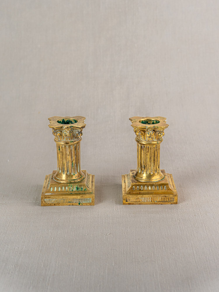 Pair of antique French gilded brass candlesticks, Louis XVI style, with column designs and square bases on light gray surface.