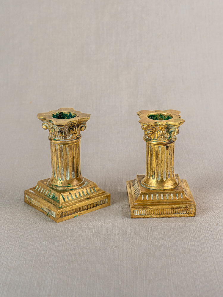 Antique French Louis XVI brass candlesticks with classical columns and square bases on a neutral textured background.