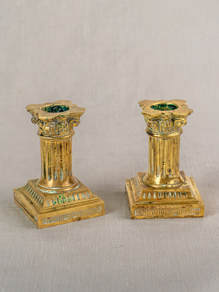 Antique French Louis XVI gilded brass candlesticks with ornate columns and square bases on light gray fabric.