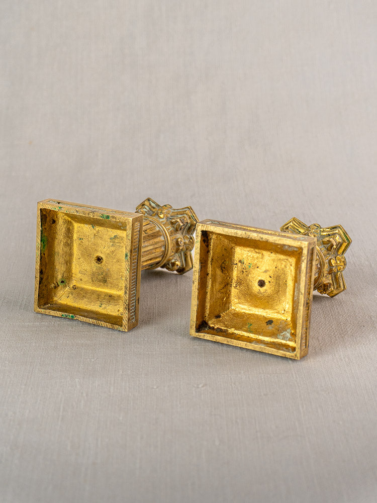 Antique French gilded brass candlesticks, Louis XVI style with ornate details and square bases, on neutral fabric.