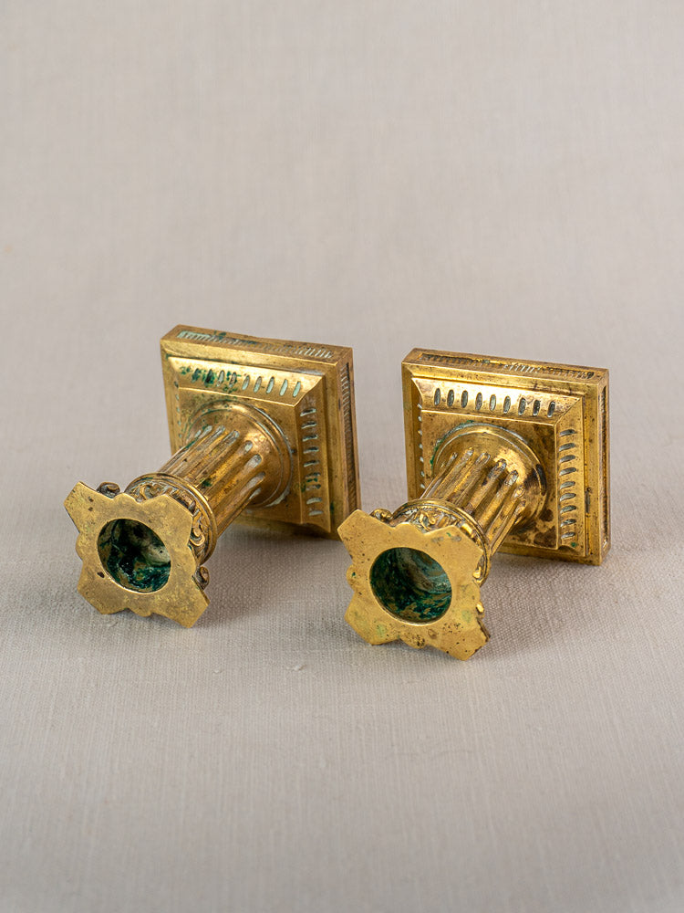 Pair of antique French Louis XVI gilded brass candlesticks with ornate tops, fluted columns, on neutral fabric background.