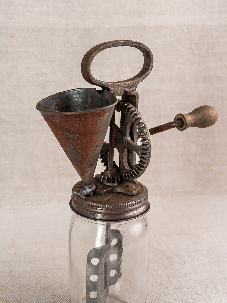 Antique French cast iron mayonnaise maker with metal funnel and gears on clear glass jar, neutral fabric backdrop.