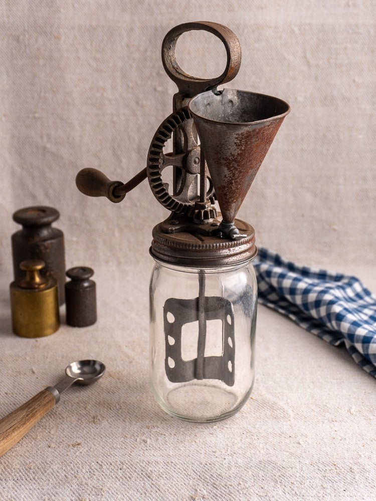 Antique French cast iron mayonnaise maker with hand-crank egg beater, glass jar, and accessories on linen.
