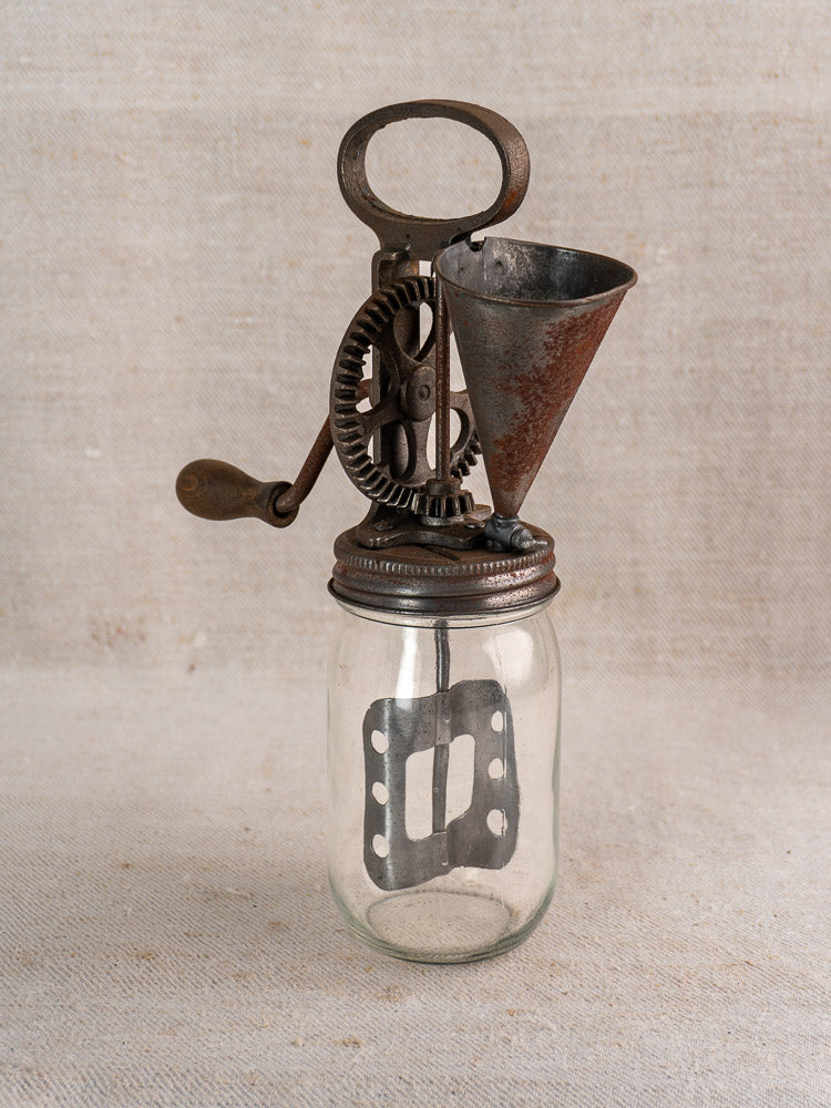 Antique French mayonnaise maker, c. 1900, with cast iron crank and glass jar, on fabric backdrop.